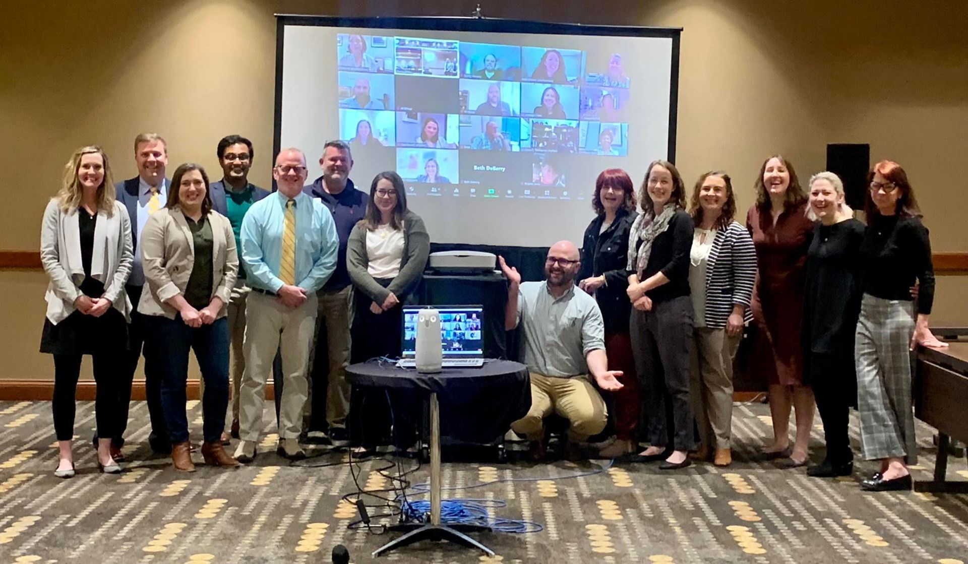 A picture of the SEMC2022 Program Committee in Rogers, Arizona. Everyone smiles at the camera and the projector screen shows the virtual attendees over Zoom. 